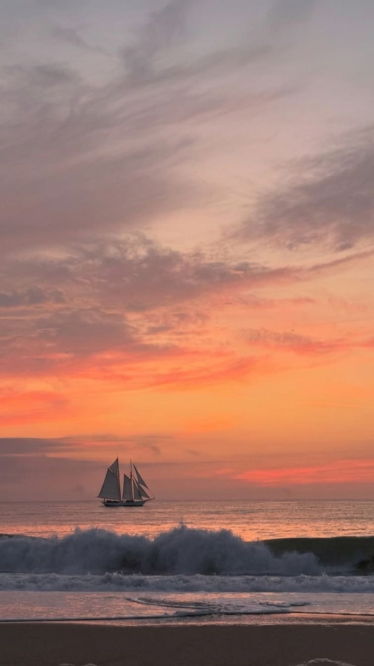 A two-masted sailboat on the ocean horizon under orange and pink clouds, with a foamy wave breaking onto a beach. AI-generated artwork.