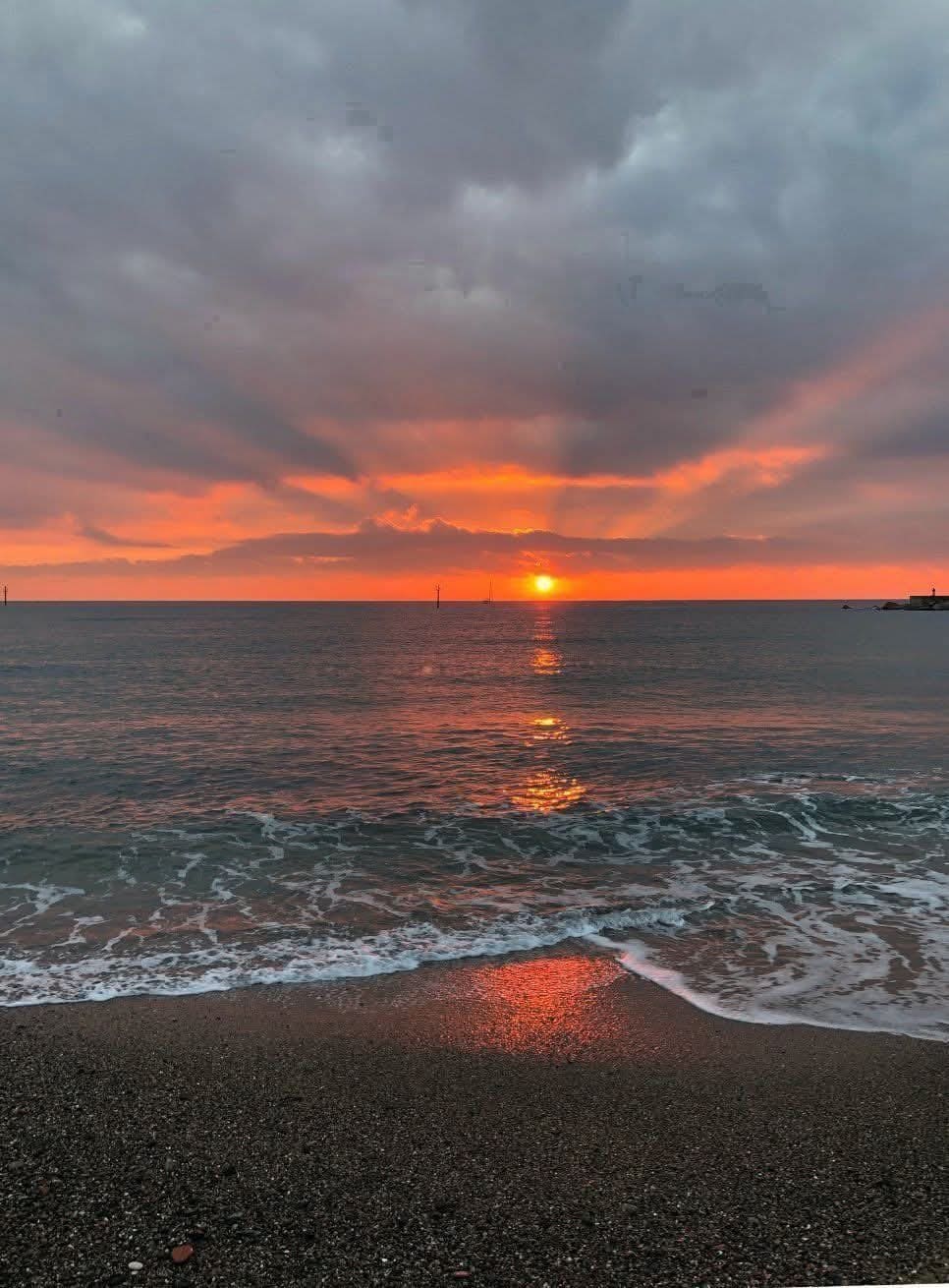 The sun on the ocean horizon sending orange rays through dark clouds, with foam on a pebbly beach. Sunset photograph.