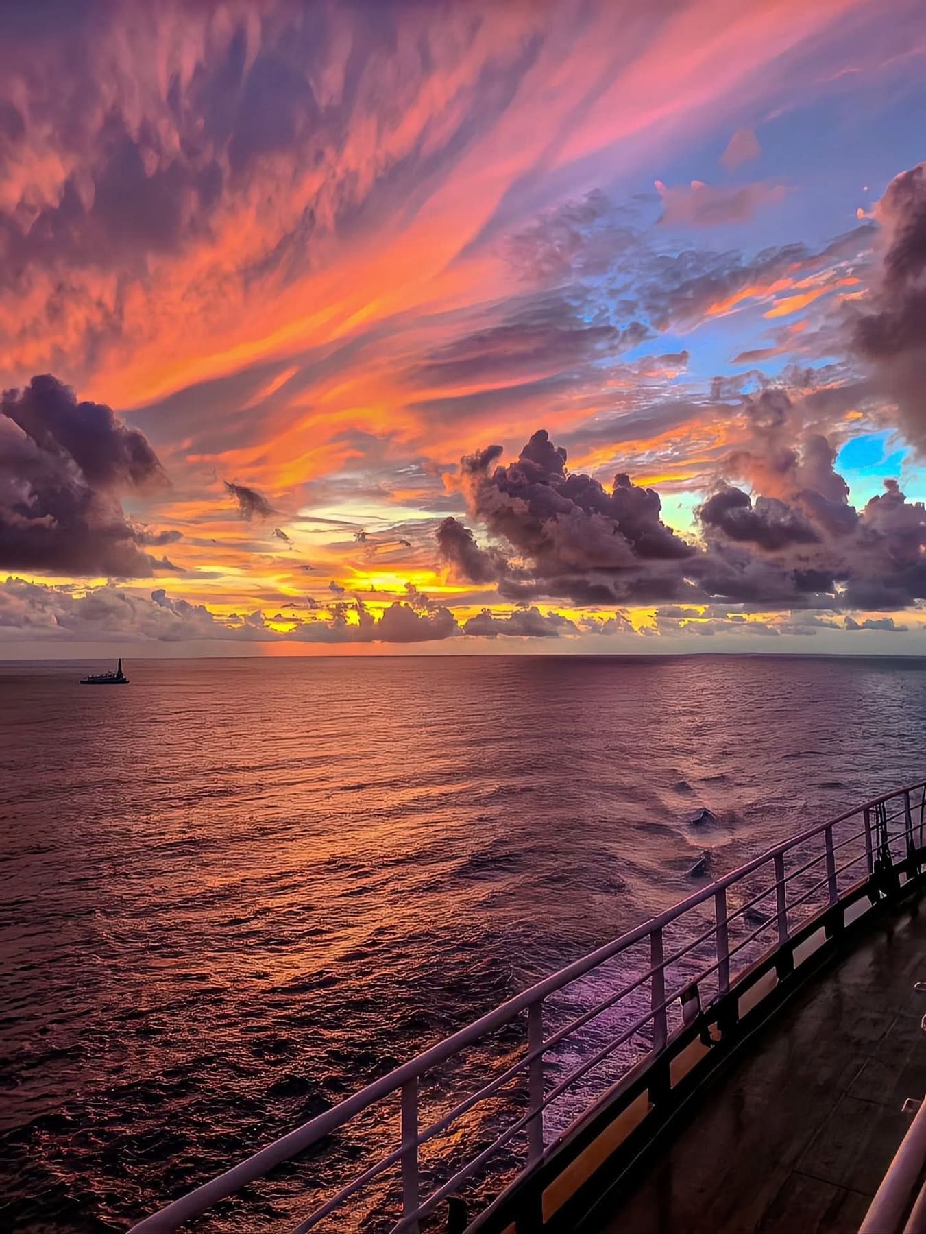 A white ship rail frames a pink, orange, and purple sunset over a dark ocean. Sunset photograph.
